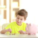A young boy in a bright yellow shirt is sitting at a table, learning to stack coins as part of teaching kids about money. Beside him, a pink piggy bank stands ready. The softly blurred background hints at a cozy indoor setting. | MONEY6X