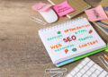 A desk with a notepad displaying colorful SEO-related words like "Web," "Content," and "SEO Things to Fix," surrounded by paper clips, sticky notes, a mouse, and a keyboard. | MONEY6X