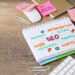 A desk with a notepad displaying colorful SEO-related words like "Web," "Content," and "SEO Things to Fix," surrounded by paper clips, sticky notes, a mouse, and a keyboard. | MONEY6X