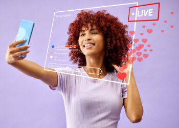A woman with curly red hair takes a selfie with her smartphone, aiming to go viral on social media. Virtual icons like "LIVE," hearts, and comments float in front of her against a vibrant purple background. | MONEY6X