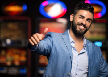 A smiling man with a beard, wearing a light blue suit, gives a thumbs-up gesture. Bright, colorful slot machines are blurred in the background, hinting at a casino setting where he might be ready to share his Slot Tips. | MONEY6X