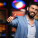 A smiling man with a beard, wearing a light blue suit, gives a thumbs-up gesture. Bright, colorful slot machines are blurred in the background, hinting at a casino setting where he might be ready to share his Slot Tips. | MONEY6X