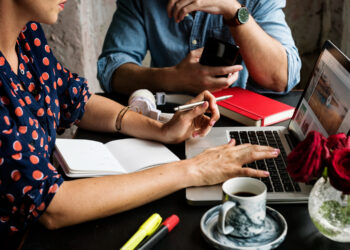 Two people work together at a table, discussing if a website is essential for an indie author. One types on a laptop while holding a pen; the other sips coffee. Headphones, highlighters, and red roses complete the creative scene. | MONEY6X