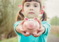 A young girl with pigtails and red polka dot ribbons holds a pink piggy bank towards the camera, standing on a blurred outdoor path—a picture of innocence in the complex world of child custody. She wears a turquoise shirt and has a calm expression. | MONEY6X
