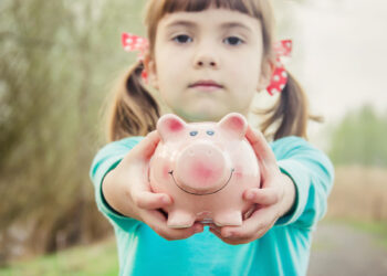 A young girl with pigtails and red polka dot ribbons holds a pink piggy bank towards the camera, standing on a blurred outdoor path—a picture of innocence in the complex world of child custody. She wears a turquoise shirt and has a calm expression. | MONEY6X