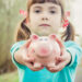 A young girl with pigtails and red polka dot ribbons holds a pink piggy bank towards the camera, standing on a blurred outdoor path—a picture of innocence in the complex world of child custody. She wears a turquoise shirt and has a calm expression. | MONEY6X