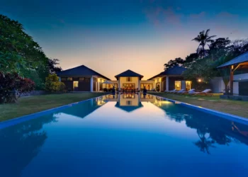 Luxurious villa in Bali at dusk with a long, illuminated pool reflecting the vibrant evening sky. The villa comprises multiple buildings, surrounded by Indonesian trees and ambient lighting, creating a serene and inviting atmosphere. | MONEY6X