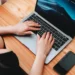 A person types on a MacBook Pro at a wooden desk, with a smartphone placed beside the laptop. The screen of the MacBook Pro displays a dark, moody wallpaper. The photo is taken from above. | MONEY6X