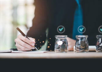 A person in a suit writes in a notebook at a desk with three glass jars filled with coins in the foreground, each jar marked with a blue checkmark, reflecting smart money management and awareness of Payday Loan Regulations. | MONEY6X