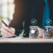 A person in a suit writes in a notebook at a desk with three glass jars filled with coins in the foreground, each jar marked with a blue checkmark, reflecting smart money management and awareness of Payday Loan Regulations. | MONEY6X