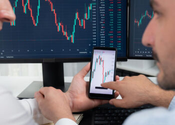 Two men analyze stock charts on computer monitors and a smartphone, discussing financial data, trends, and Pre-IPO Stocks displayed on the screens. | MONEY6X