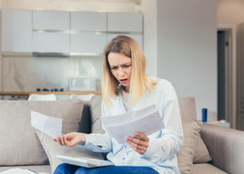 A woman sits on a couch holding papers and looking at them with a worried expression, her laptop open in front of her in a modern home setting, as she searches for ways to reduce your tax bill. | MONEY6X