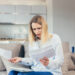 A woman sits on a couch holding papers and looking at them with a worried expression, her laptop open in front of her in a modern home setting, as she searches for ways to reduce your tax bill. | MONEY6X