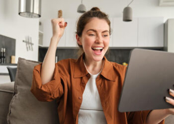 A young woman sitting on a couch in a modern living room smiles and raises her fist in excitement while looking at a tablet, thrilled to discover new ways to earn money from home. | MONEY6X
