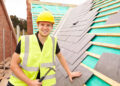 A smiling construction worker in a yellow hard hat and reflective vest holds a hammer while installing slate tiles on a roof under construction. | MONEY6X