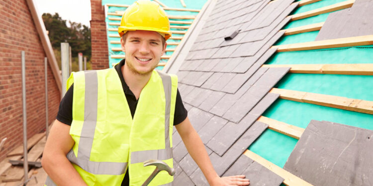 A smiling construction worker in a yellow hard hat and reflective vest holds a hammer while installing slate tiles on a roof under construction. | MONEY6X