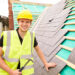 A smiling construction worker in a yellow hard hat and reflective vest holds a hammer while installing slate tiles on a roof under construction. | MONEY6X
