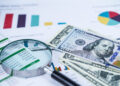 A magnifying glass, spreadsheets, colorful charts, and several U.S. $100 bills on a desk, representing financial analysis, business reports, money management, and personal wealth creation. | MONEY6X