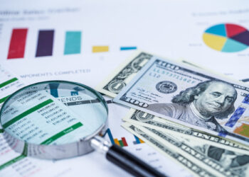 A magnifying glass, spreadsheets, colorful charts, and several U.S. $100 bills on a desk, representing financial analysis, business reports, money management, and personal wealth creation. | MONEY6X