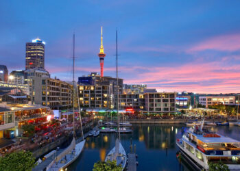 A lively waterfront marina at sunset in Auckland with boats docked, modern buildings lit up, and the Sky Tower illuminated in the background under a colorful sky. | MONEY6X