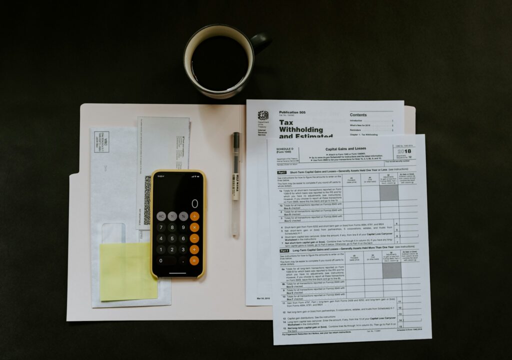 A folder, tax envelopes, sticky notes, a smartphone with a calculator app open, a pen, a cup of black coffee, and IRS tax documents are arranged on a dark surface. | MONEY6X