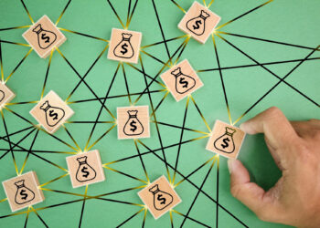 A hand picks up a wooden tile with a money bag icon among several similar tiles, all connected by black lines on a green background, symbolizing the 50/30/20 Rule to Money Management within a financial network. | MONEY6X