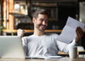 A man sitting at a table with a laptop and coffee cup smiles and raises his fist in excitement while reading a document, showing the satisfaction of getting more from every invoice you send as a freelancer. | MONEY6X