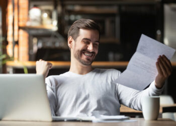 A man sitting at a table with a laptop and coffee cup smiles and raises his fist in excitement while reading a document, showing the satisfaction of getting more from every invoice you send as a freelancer. | MONEY6X