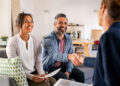 A smiling couple sits on a couch, with the man shaking hands with a professional woman across from them. The woman holds papers, and they appear to be in a bright, modern living room. | MONEY6X