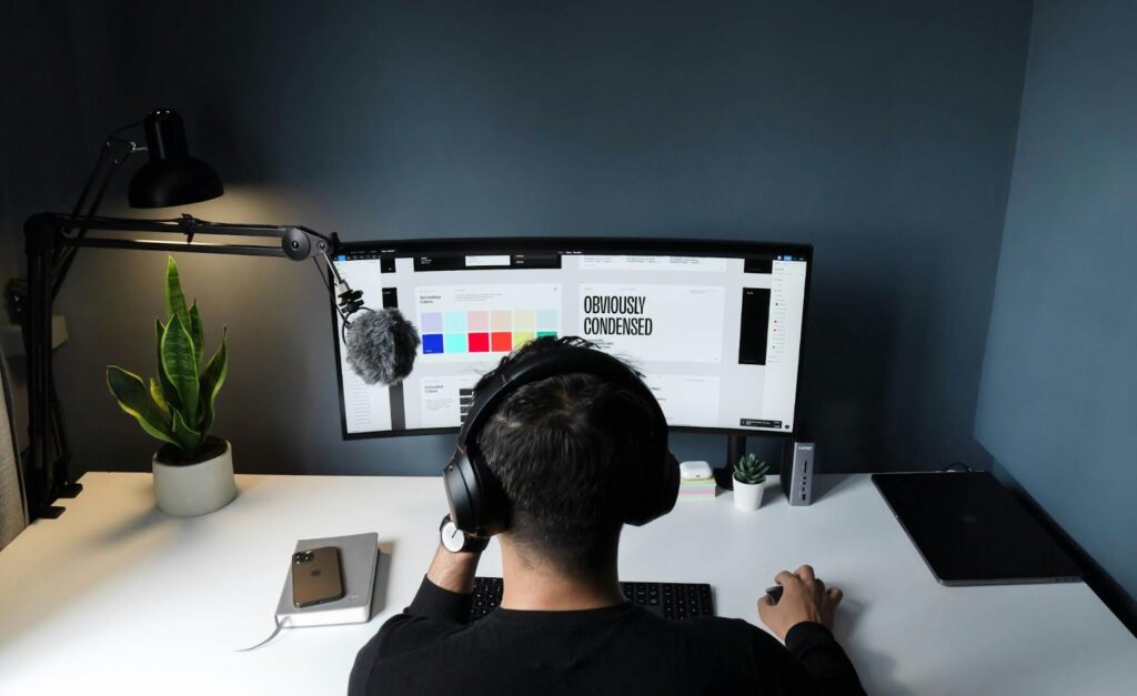 A Freelancer wearing headphones sits at a desk with a large curved monitor displaying design software and an invoice, alongside a microphone, potted plant, smartphone, and closed laptop, in a modern, minimalistic workspace. | MONEY6X
