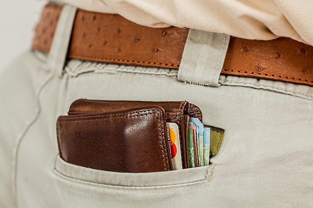 A brown leather wallet filled with cards and cash sticks out of the back pocket of light-colored pants worn with a brown belt. | MONEY6X