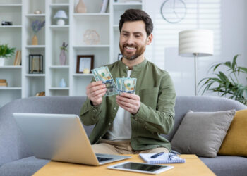 A smiling man sits on a couch in a modern living room, holding several hundred-dollar bills—his laptop, tablet, and notepad on the coffee table hint at recent online games or success. | MONEY6X