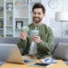 A smiling man sits on a couch in a modern living room, holding several hundred-dollar bills—his laptop, tablet, and notepad on the coffee table hint at recent online games or success. | MONEY6X