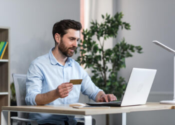 A man sits at a desk holding a credit card in one hand and using a laptop with the other, suggesting online shopping, banking, or managing an online business. The background includes a plant, bookshelves, and a modern desk lamp. | MONEY6X