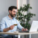 A man sits at a desk holding a credit card in one hand and using a laptop with the other, suggesting online shopping, banking, or managing an online business. The background includes a plant, bookshelves, and a modern desk lamp. | MONEY6X