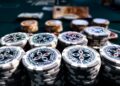 Stacks of poker chips in various colors are arranged on a green felt table, with blurred chips and cards in the background. The focus is on the front chips, labeled "Ultimate Poker Chip. | MONEY6X