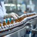 A worker in gloves inspects brown glass bottles with white caps on a conveyor belt in a pharmaceutical lab.