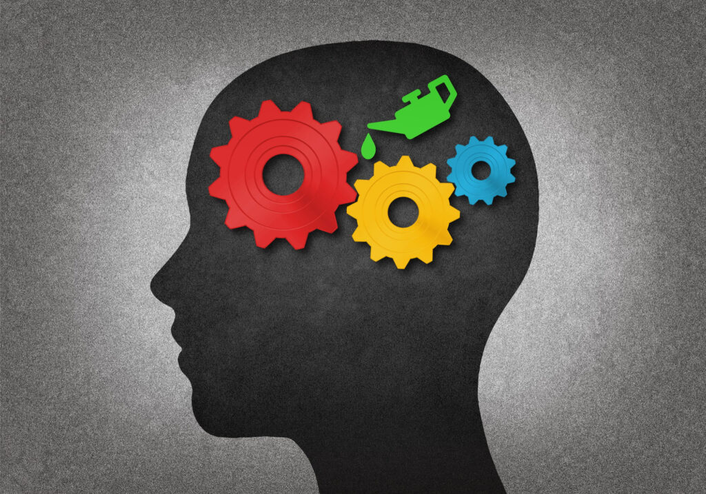 A silhouette of a human head with three colored gears (red, yellow, blue) inside the brain area and a green oil can above, suggesting the idea of maintaining or fueling the mind. | MONEY6X