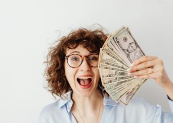 Smiling woman with glasses holding a fan of US dollar bills in her hand against a white background.