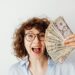 Smiling woman with glasses holding a fan of US dollar bills in her hand against a white background.