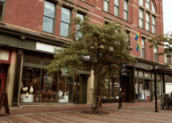 A brick building with large windows houses shops at street level. A tree stands on the sidewalk, and a rainbow flag hangs above one store. Clothing racks and displays are visible outside the shops. | MONEY6X