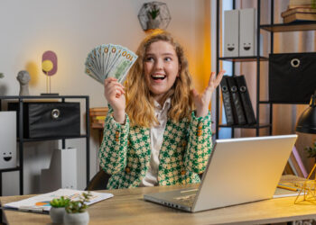 A smiling woman sitting at a desk with a laptop holds a fan of dollar bills in one hand and raises her other hand in excitement, suggesting financial success or a windfall in a modern office setting. | MONEY6X