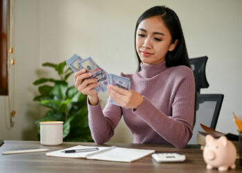 A woman in a purple sweater sits at a desk, counting US dollar bills. On the desk are a notebook, calculator, pencil holder, coffee mug, and a piggy bank. A plant is visible in the background. | MONEY6X