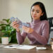 A woman in a purple sweater sits at a desk, counting US dollar bills. On the desk are a notebook, calculator, pencil holder, coffee mug, and a piggy bank. A plant is visible in the background. | MONEY6X