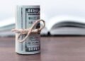 A roll of U.S. hundred dollar bills tied with twine sits on a wooden surface with an open book blurred in the background. | MONEY6X