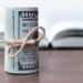 A roll of U.S. hundred dollar bills tied with twine sits on a wooden surface with an open book blurred in the background. | MONEY6X