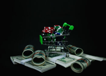 A small shopping cart filled with poker chips sits on top of several stacks and rolls of U.S. hundred dollar bills against a black background. | MONEY6X