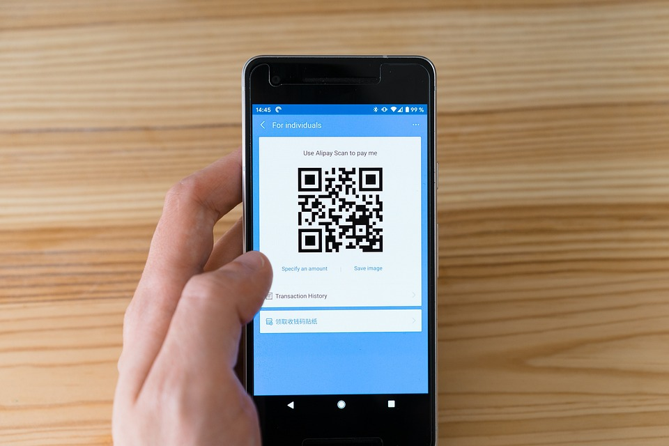 A hand holding a smartphone displaying a payment app with a QR code on the screen, ready to be scanned for digital payment. The phone is above a light wooden surface. | MONEY6X