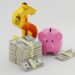 A pink piggy bank, a large gold dollar sign, and several stacks of U.S. hundred-dollar bills are arranged on a plain white background, symbolizing saving and financial growth. | MONEY6X