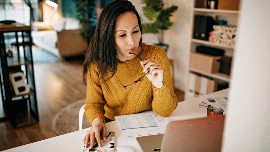 A woman in a yellow sweater sits at a desk, holding glasses and looking at a laptop. Papers, a mug, and a notebook are on the desk. She appears focused and thoughtful, working in a cozy, well-lit home environment. | MONEY6X
